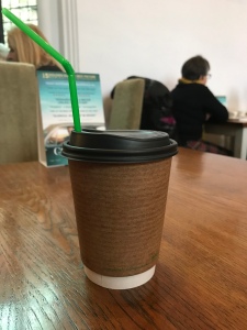 A photo of my compostable cup with the green biodegradable straw. The cup is on a wooden table. You can see other customers in the distance. 