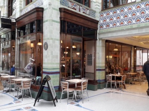 A photo taken outside Marmalades café yet still inside Royal Arcade. There is seating outside the café. Big windows give you a glimpse inside. There is Art Nouveau tile work on the architecture. 
