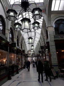Looking down the long shopping area of Royal Arcade Norwich. There is a curved glass roof, and black metalwork ornate light fittings hanging down like lamps. 