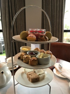 The afternoon tea stand in the centre of our table with a teapot to the left. There are colourful desserts on the top layer, scones in the middle, and sandwiches at the bottom.