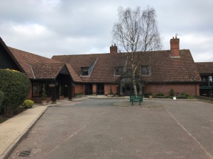 A photo taken wheeling towards the main entrance at Barnham Broom Country Club. It is a long l shaped brick building. The sky is grey with clouds. 