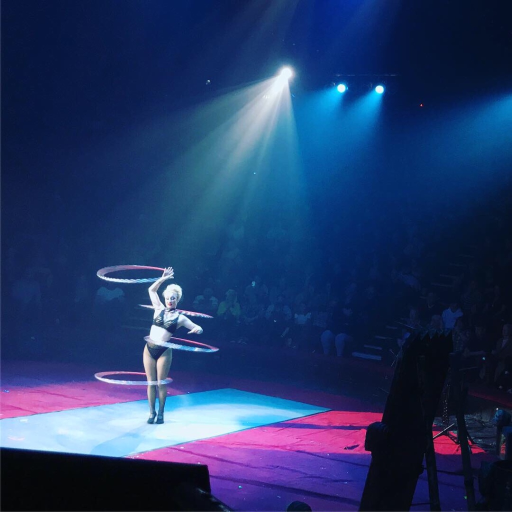 A lady with short white hair and a black bikini style clothing is hula hooping with multiple hoops on her body and arm. 
