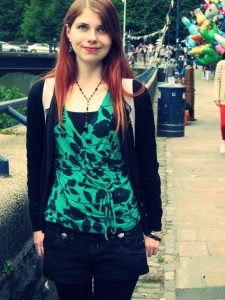 A photo of Caz standing in the street by a river. She has red hair, a green and black top, black cardigan, denim shorts and thick black tights. You can not tell Caz has any disability. 