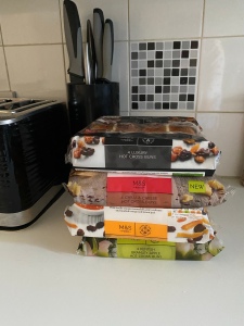 A pile of hot cross buns on my kitchen side in packets. The flavours are, luxury fruit, chilli and cheese, orange marmalade, and apple. The kitchen counter is white, the tiles and black and white, plus you can see a black toaster and knife block. 